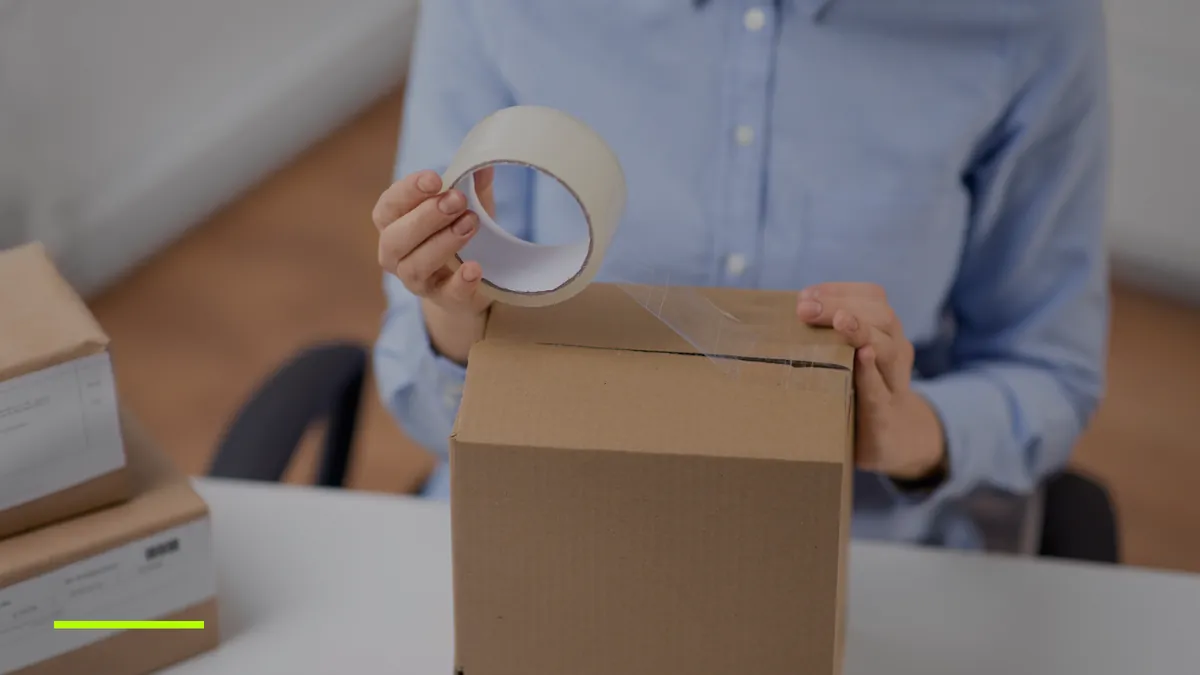 Mâini care sigilează cu bandă adezivă un colet simplu din carton